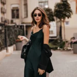 fashionable-pale-brunette-long-green-dress-black-jacket-sunglasses-standing-street-during-daytime-against-wall-light-city-building_197531-24468