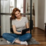 satisfied-girl-jeans-striped-top-sits-floor-living-room-works-laptop-with-smile_197531-14785