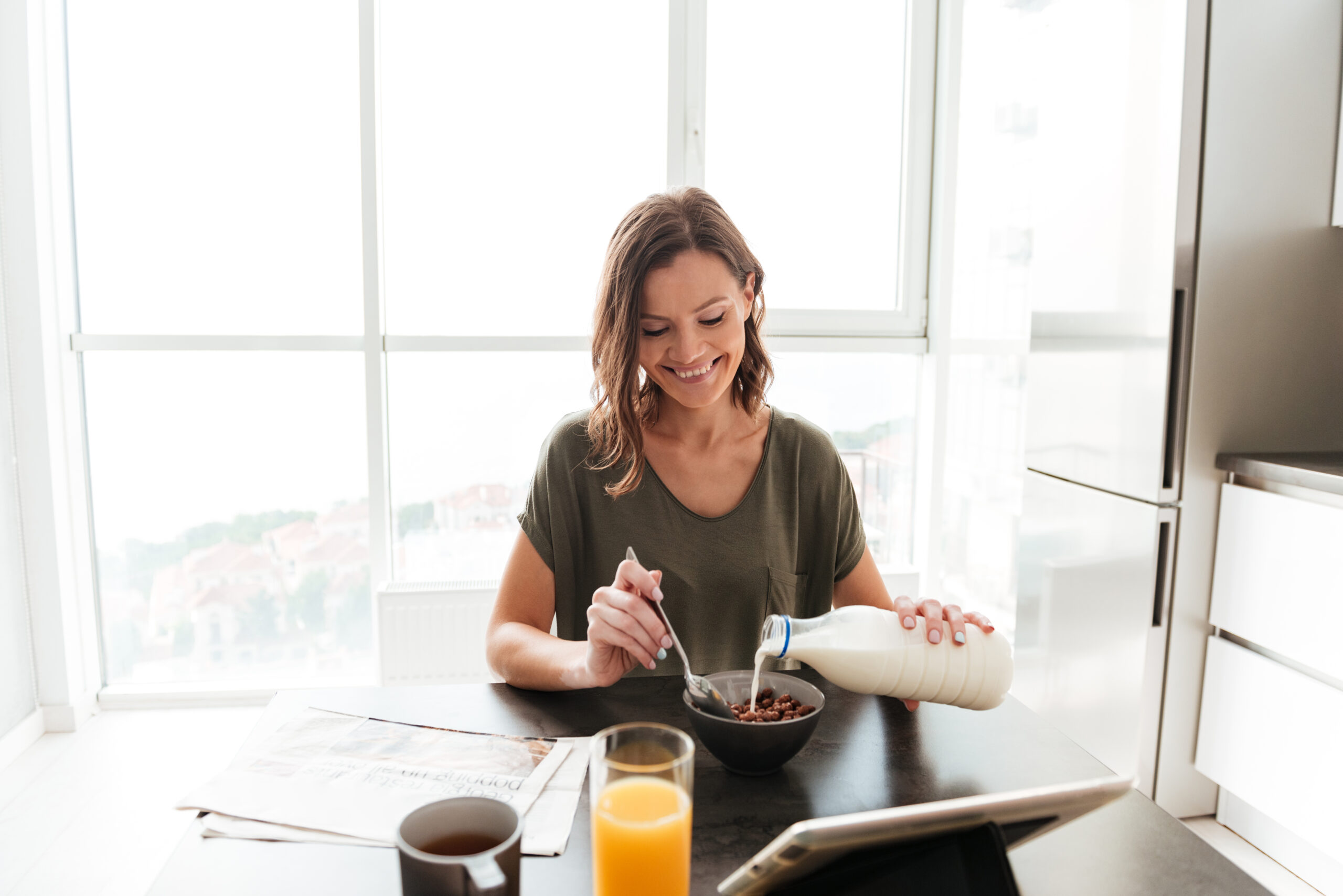 Pleased casual woman eating by the table on kitchen and looking at the tablet computer